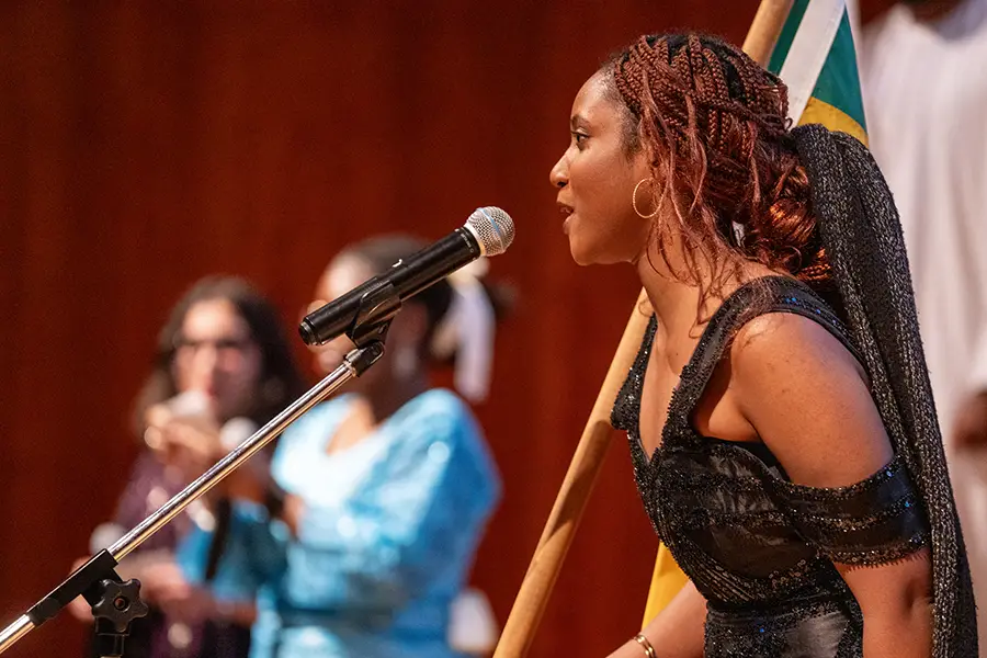 A student stands at a microphone on stage in traditional dress holding a country flag.
