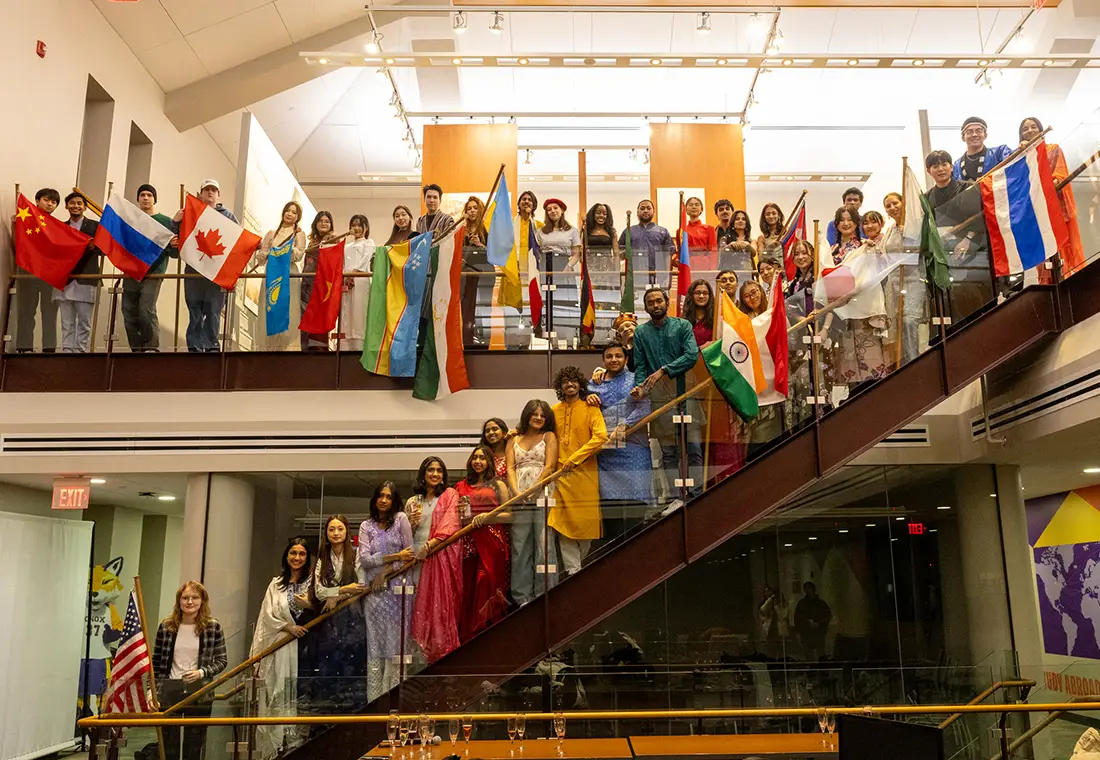 A large group of students in traditional dress hold their countries flag in a line going up the stairs and long the second level of Alumni Hall on Knox College campus.
