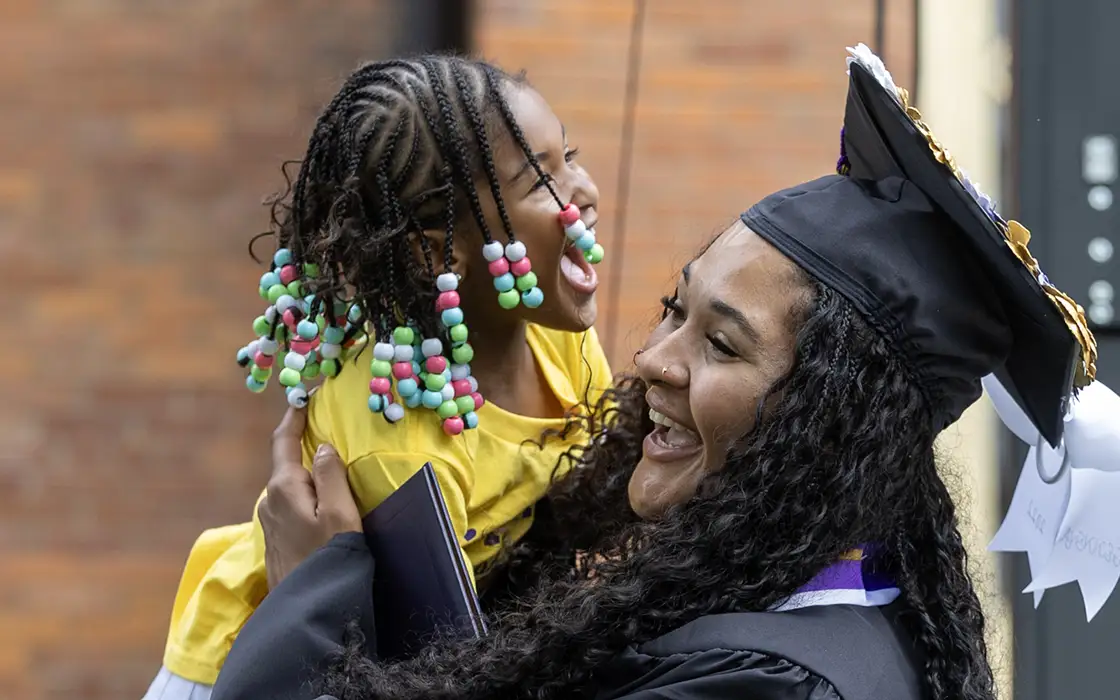 A graduate holds a young child, along with her diploma. The child laughs joyfully. 