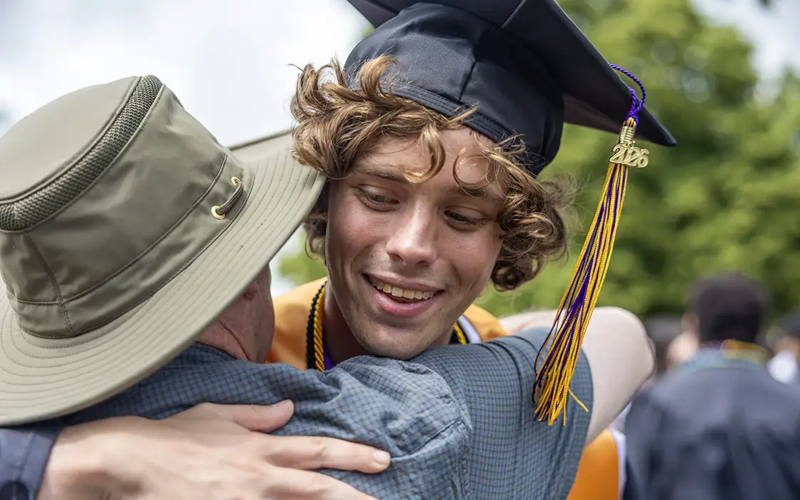 A graduate leans in to hug an individual. The graduate's 2026 tassel swings forward. 