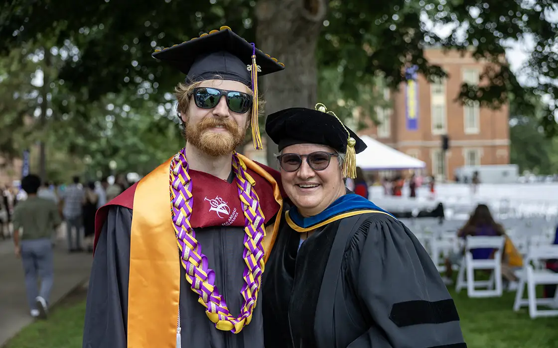 Two individuals stand on the South Lawn of Old Main, wearing academic regalia and smiling at the camera. 