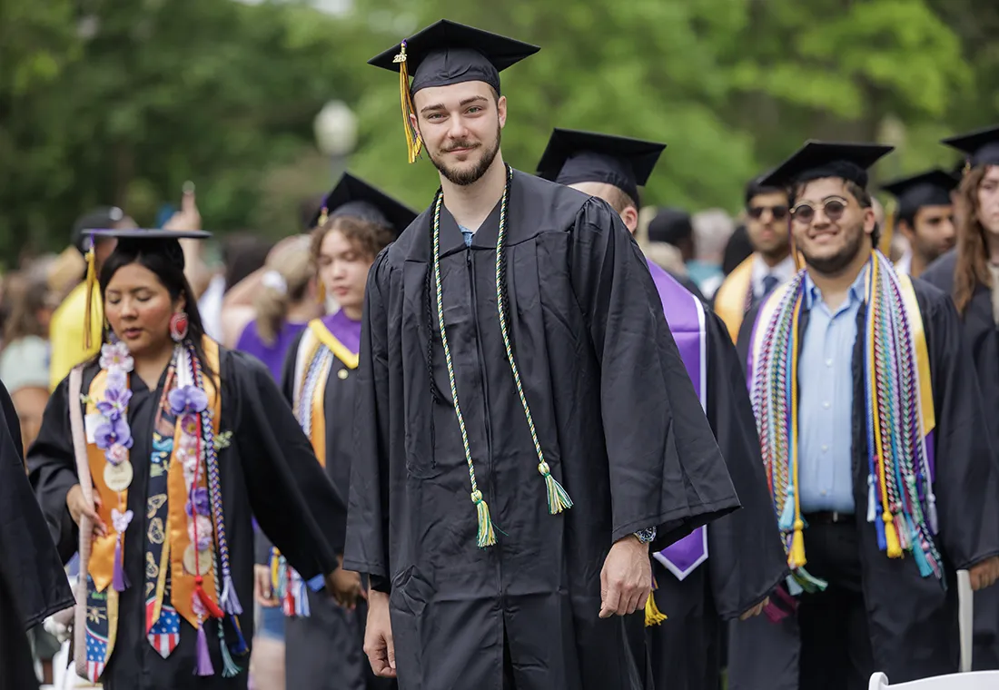 A graduate walks, smiling directly at the camera. Several other graduates are walking behind him.