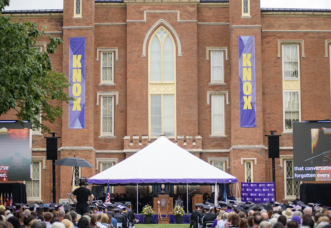 A crowd is seated in chairs on the South Lawn of Old Main; a tent is set up in front of the building, a speaker stands before a podium under the tent.