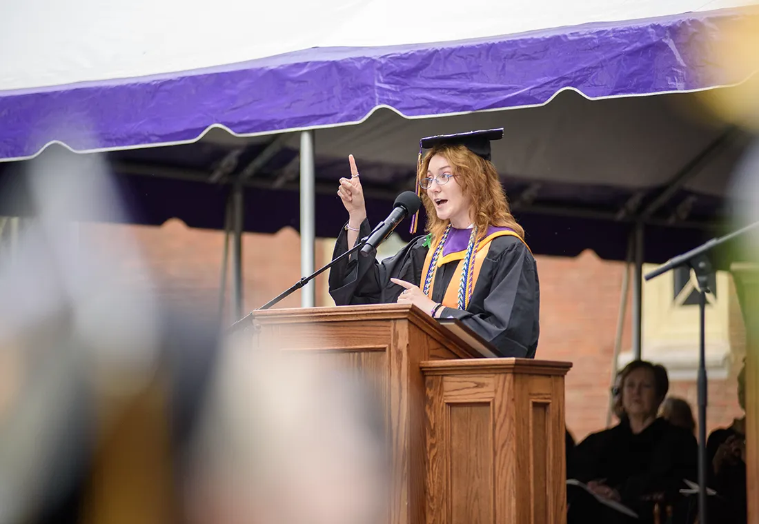 Patience Claire Wagner stands behind a podium, speaking. She wears a cap and gown, and she holds up her right index finger.