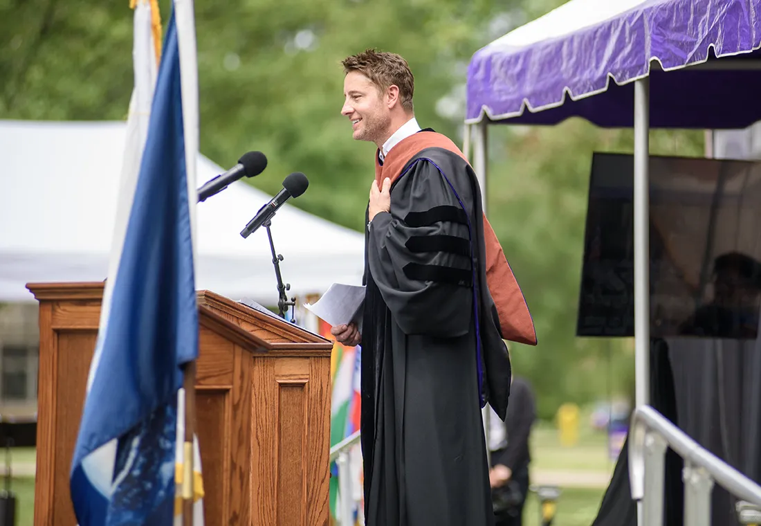 Justin Hartley stands behind a podium wearing academic regalia. He smiles, and his hand is on his chest.