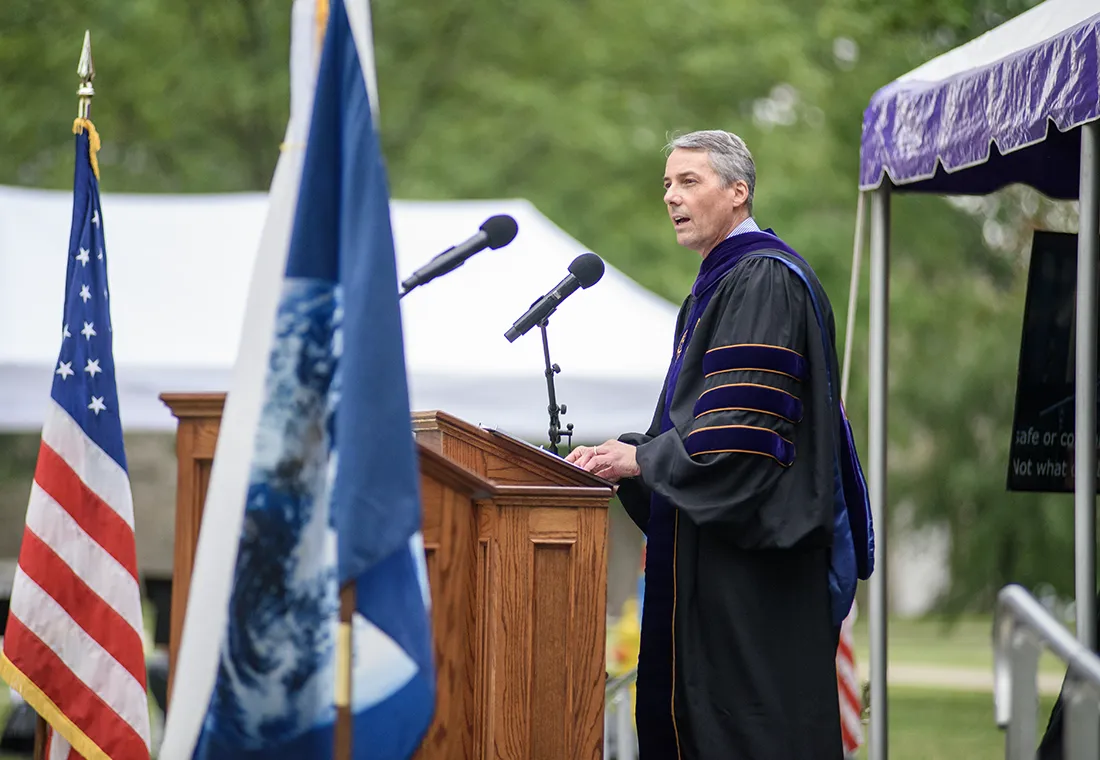 John Lawler ’88 stands at a podium, speaking. He wears academic regalia.