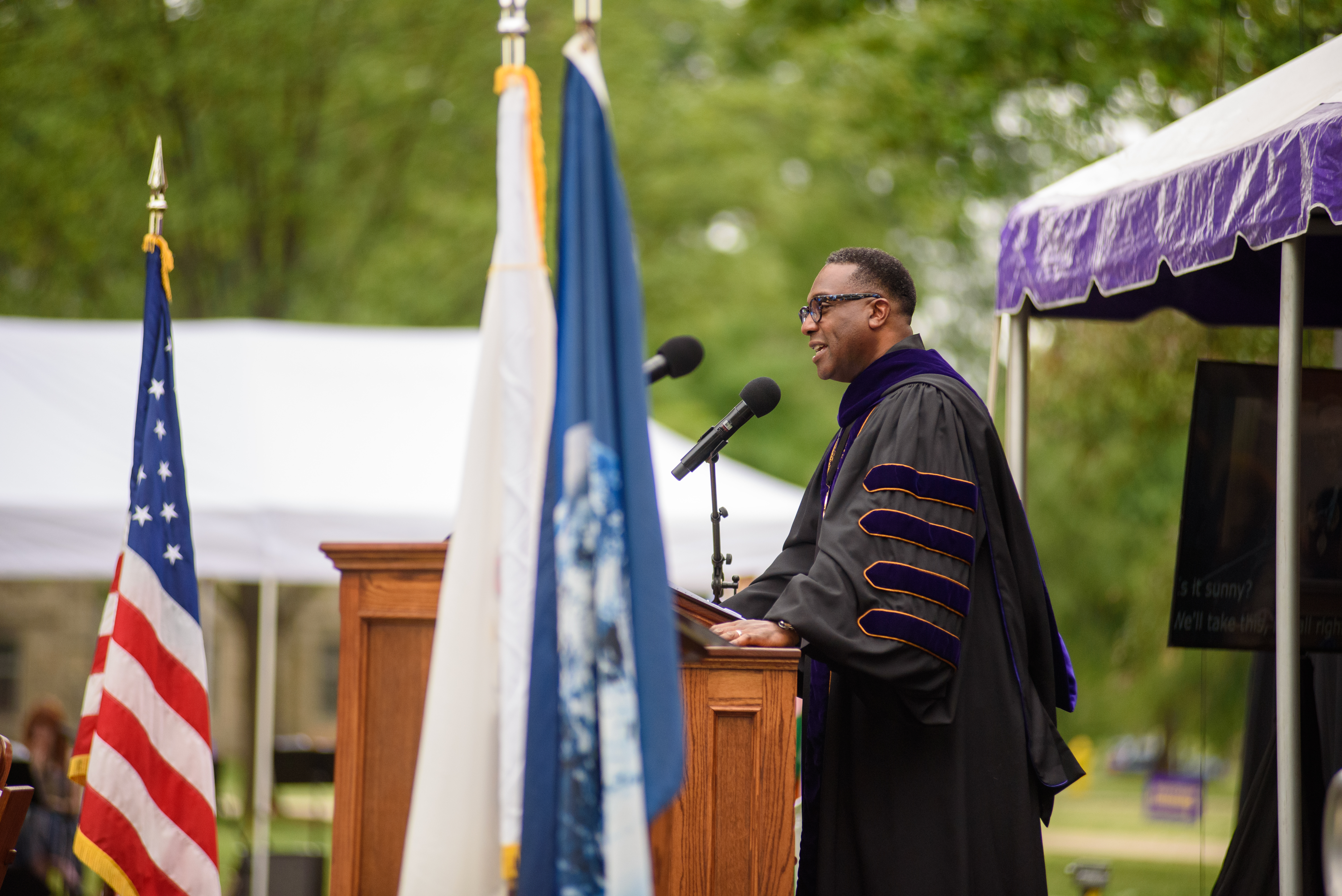 President McGadney, dressed in academic regalia, stands behind a podium on a stage and speaks.