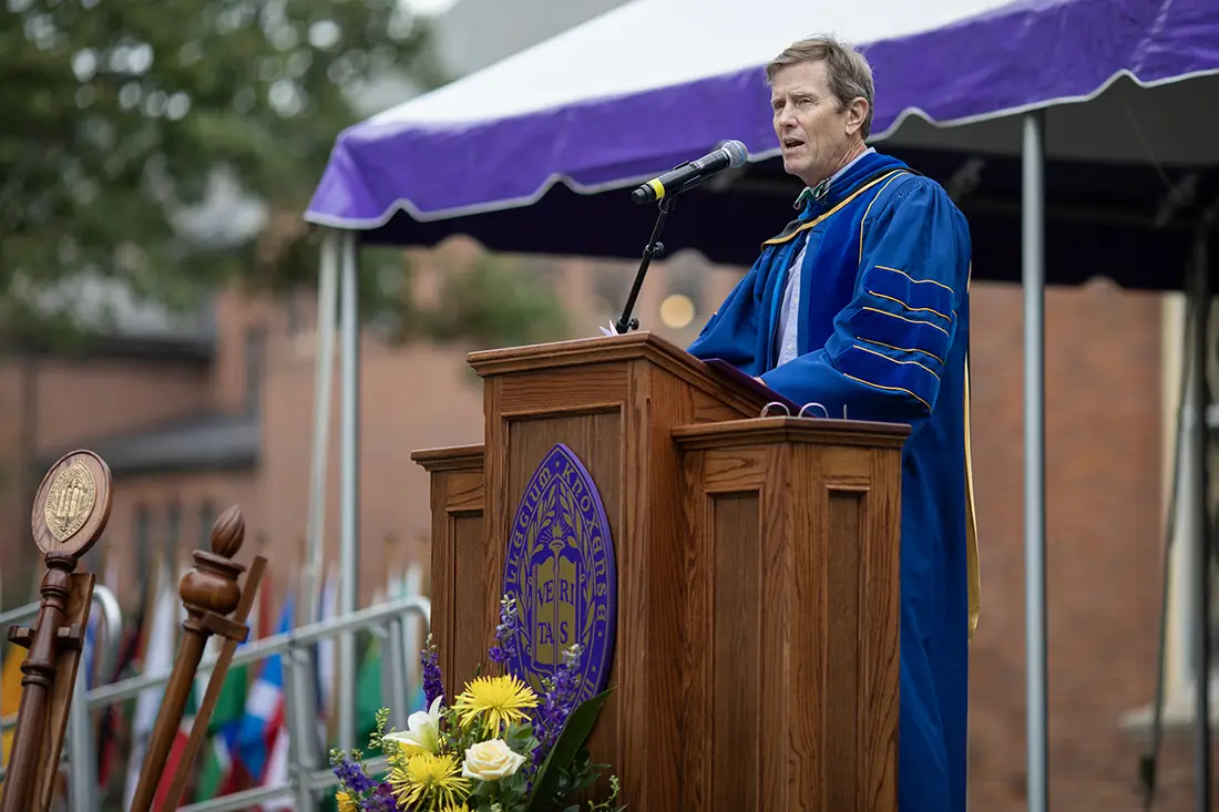 A person wearing academic regalia stands in front of a podium, speaking. The Knox seal is visible on the podium.