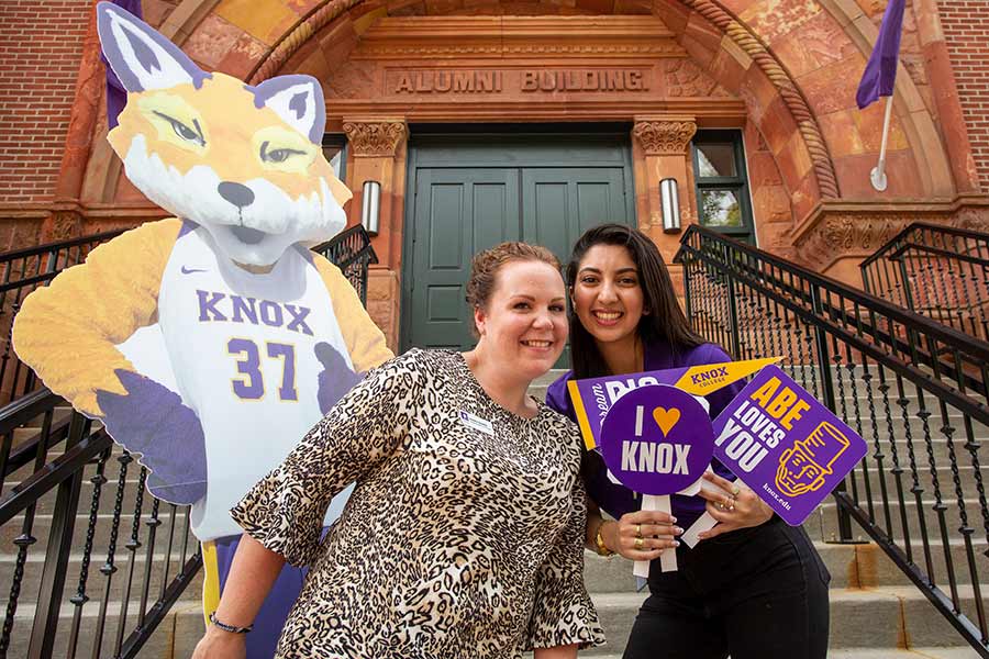 Two admission ambassadors pose for a photo with a cutout of Blaze, the Prairie Fire mascot, on the front steps of Alumni Hall.