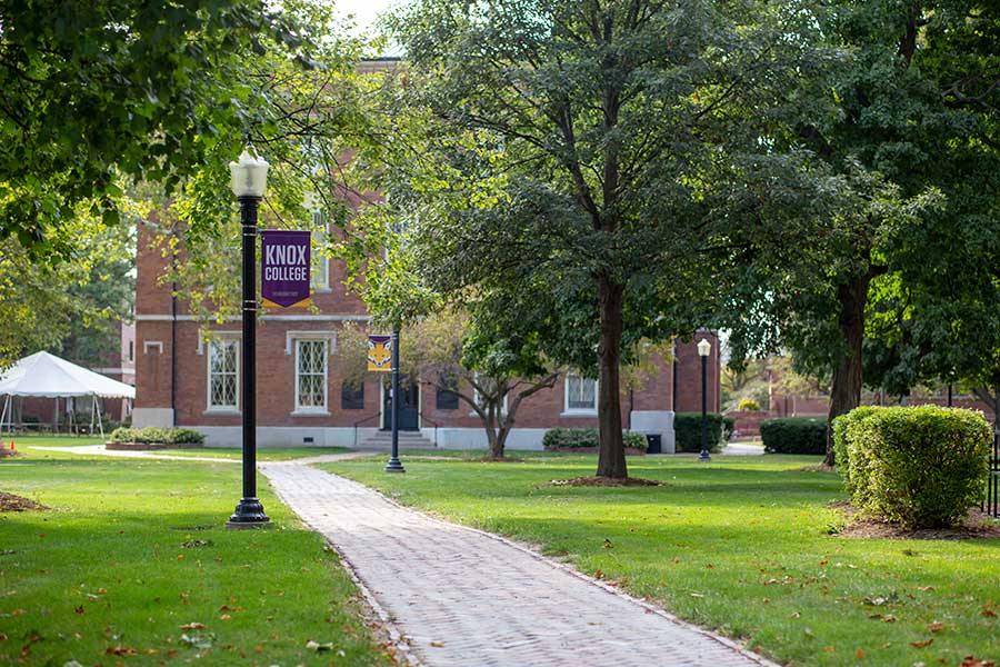 A bold purple banner reading "KNOX COLLEGE" hangs from a light pole outside Old Main on the Knox College campus.