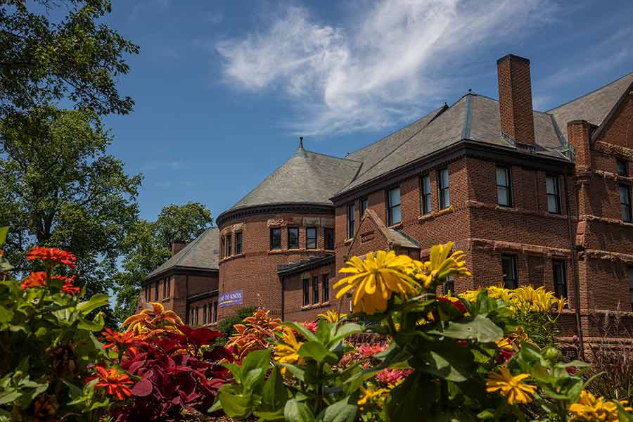 Colorful flowers in the foreground, and Alumni Hall in the background against a blue sky with a wispy cloud