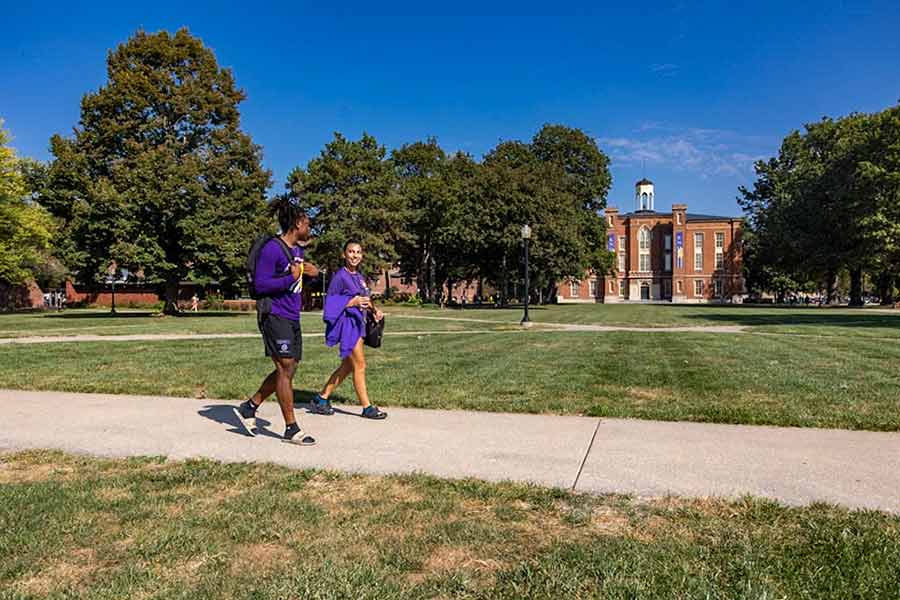 Two students walking on a sidewalk with the South Lawn and Old Main in the background against a blue sky