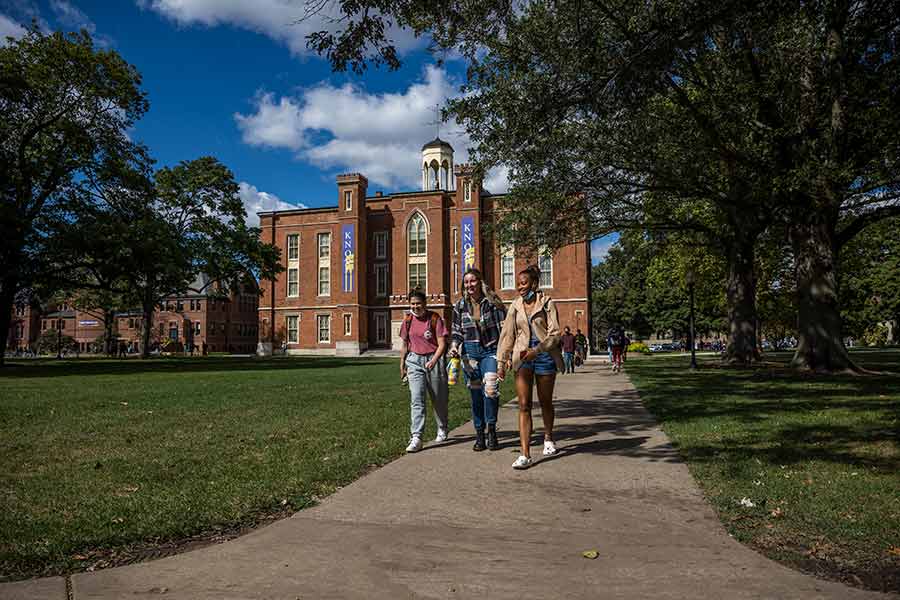 Three smiling students walk on the sidewalk with Old Main in the background