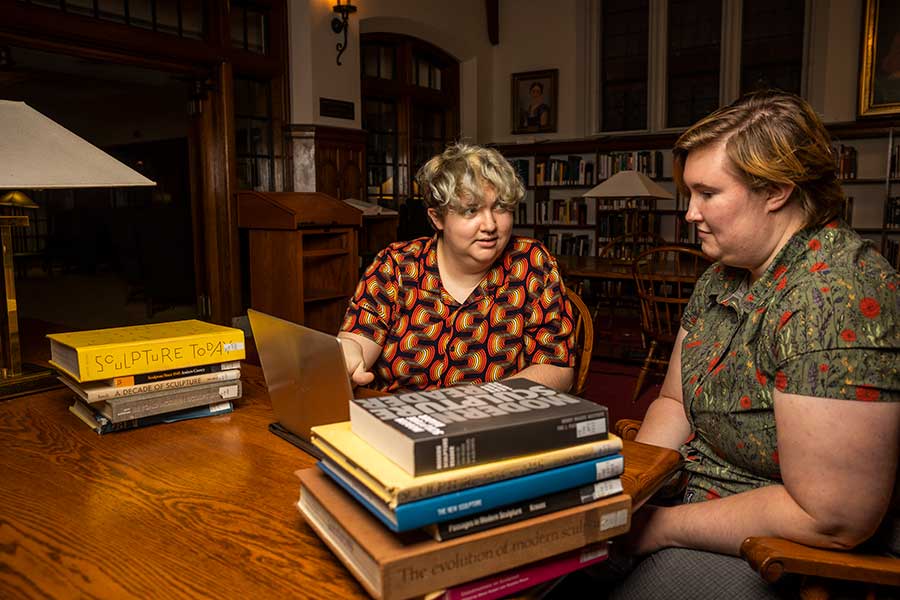 Owen, left, in red and yellow buttoned shirt, sits next to friend in green button-up with bird print in front of a wooden library table with two stacks of books on each side.
