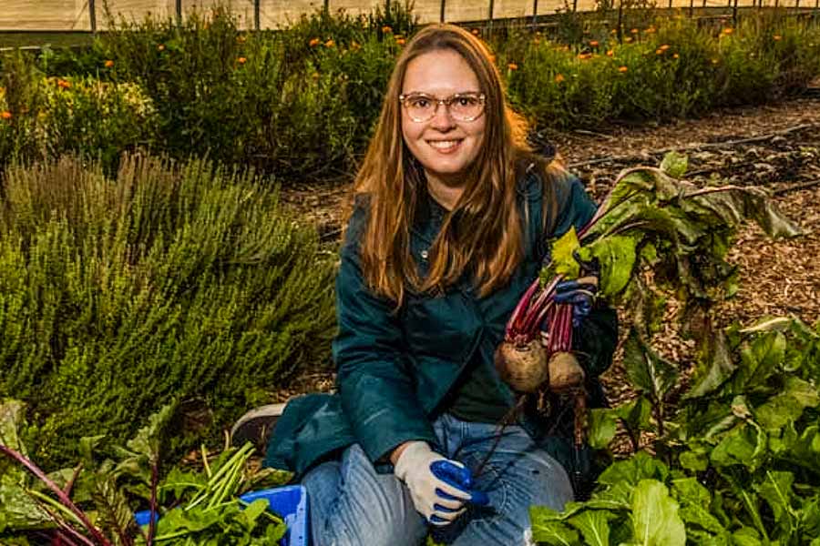 Teagan Springer, who wears a dark turquoise dress jacket, jeans, and gardening gloves, sits among green plants and cabbage holding beets in left arm.