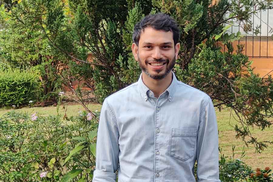 Zarir DeVitre, wearing a light blue collared shirt, stands outside near shrubbery and trees