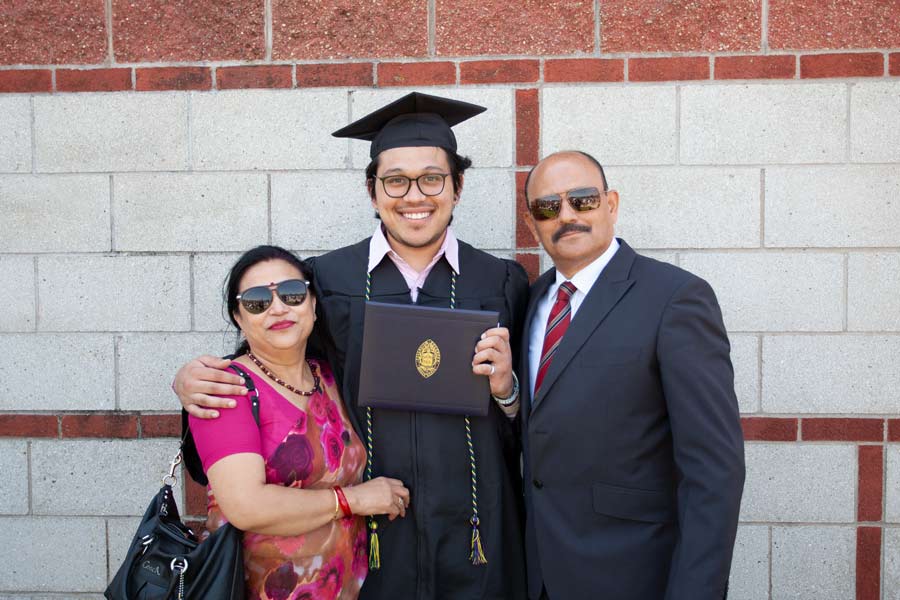 A graduating senior wearing his cap and gown and standing next to two family members outside after the Commencement ceremony.