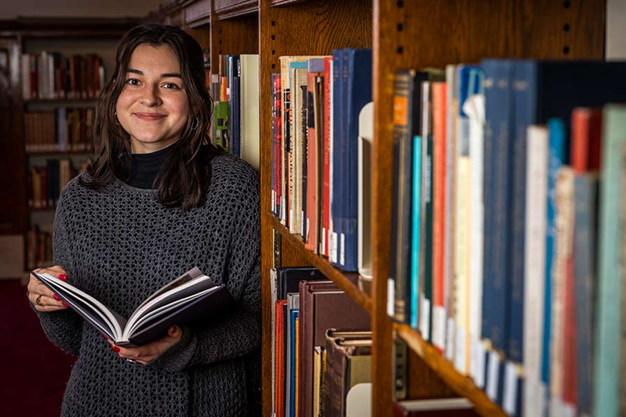 Alyx, who wears a black sweater and has shoulder length, straight, black hair holds a book and leans upon a library bookshelf.