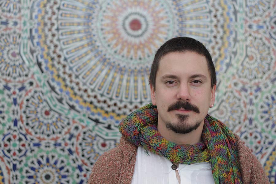 Gustavo Cortiñas, who has a mustache and a goatee, stands in front of a traditional Latin American tapestry with circular patterns with a green scarf and white shirt underneath a light brownish cardigan. 