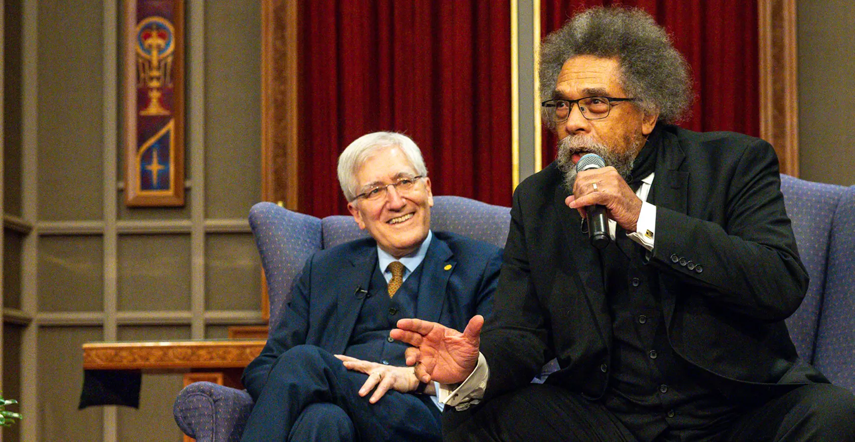 A man speaks into a microphone, sitting forward in his chair. Another man leans back in his chair and smiles as the other speaks.