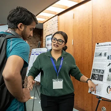 A student points to a poster on an easel as she speaks to another student.
