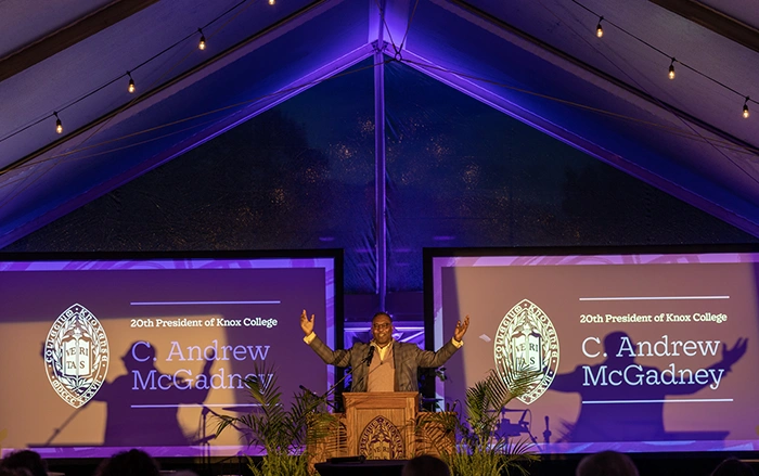 President C. Andrew McGadney stands at the front of an outdoor tent that's cast in a purple hue. His arms are spread wide as he speaks from a podium. Large screens to his right and left read: 20th President of Knox College C. Andrew McGadney.