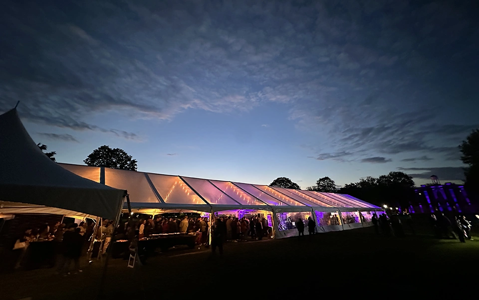 A clear, outdoor event tent set against an evening sky, lit from within in vibrant shades of purple.