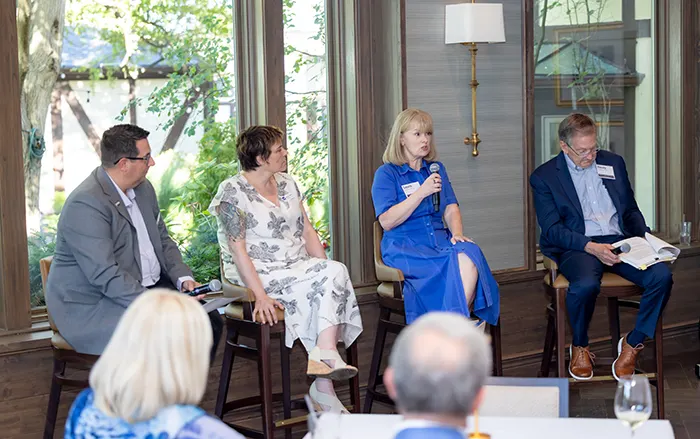 A woman holds a microphone and speaks. Three other individuals sit next to her.