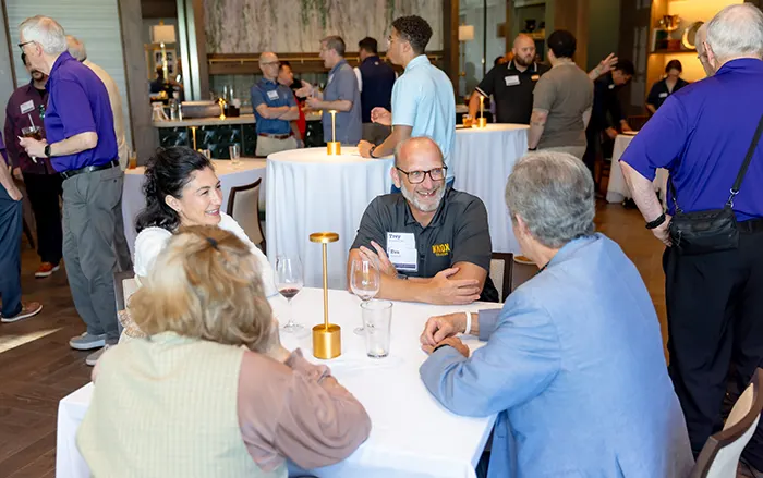 Four people sitting at a table talk and smile. Behind them, the room is full of groups of people visiting with each other.