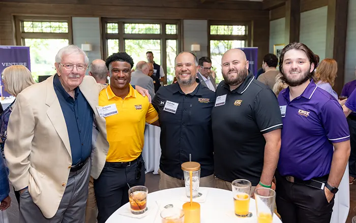 Five men with arms around each other smile at the camera. 
