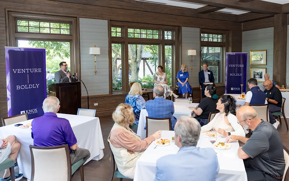 People sitting at tables look toward the front of the room at a man behind a podium with a microphone next to a purple banner that says Venture Boldly.  