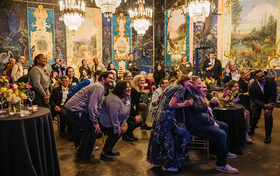 All attendees of the event smile from their tables as the speaker takes a selfie from the stage.