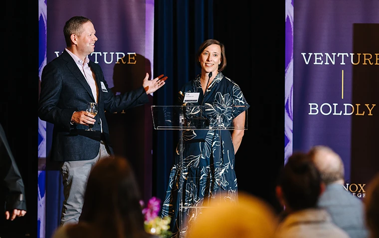 A man and woman speak on stage, flanked by purple Venture Boldly banners.