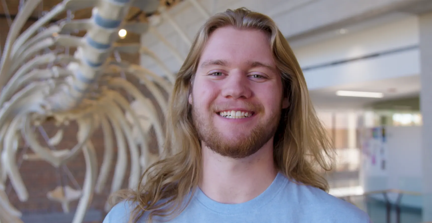 Student Iam Smith smiles at the camera inside the science and mathematics center (SMC) building in front of the whale. The whale's skeletons are visible in the background.